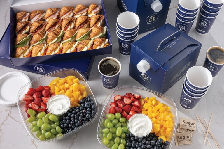Breakfast catering spread from Paris Baguette featuring trays of assorted croissant sandwiches, fresh fruit platters with strawberries, grapes, blueberries, and mango, cups of yogurt, hot coffee, and stacked paper cups arranged on a table.